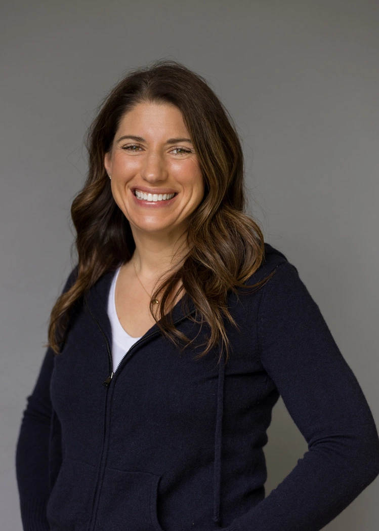 A woman smiling confidently while wearing a dark hoodie over a white shirt, standing against a gray background.